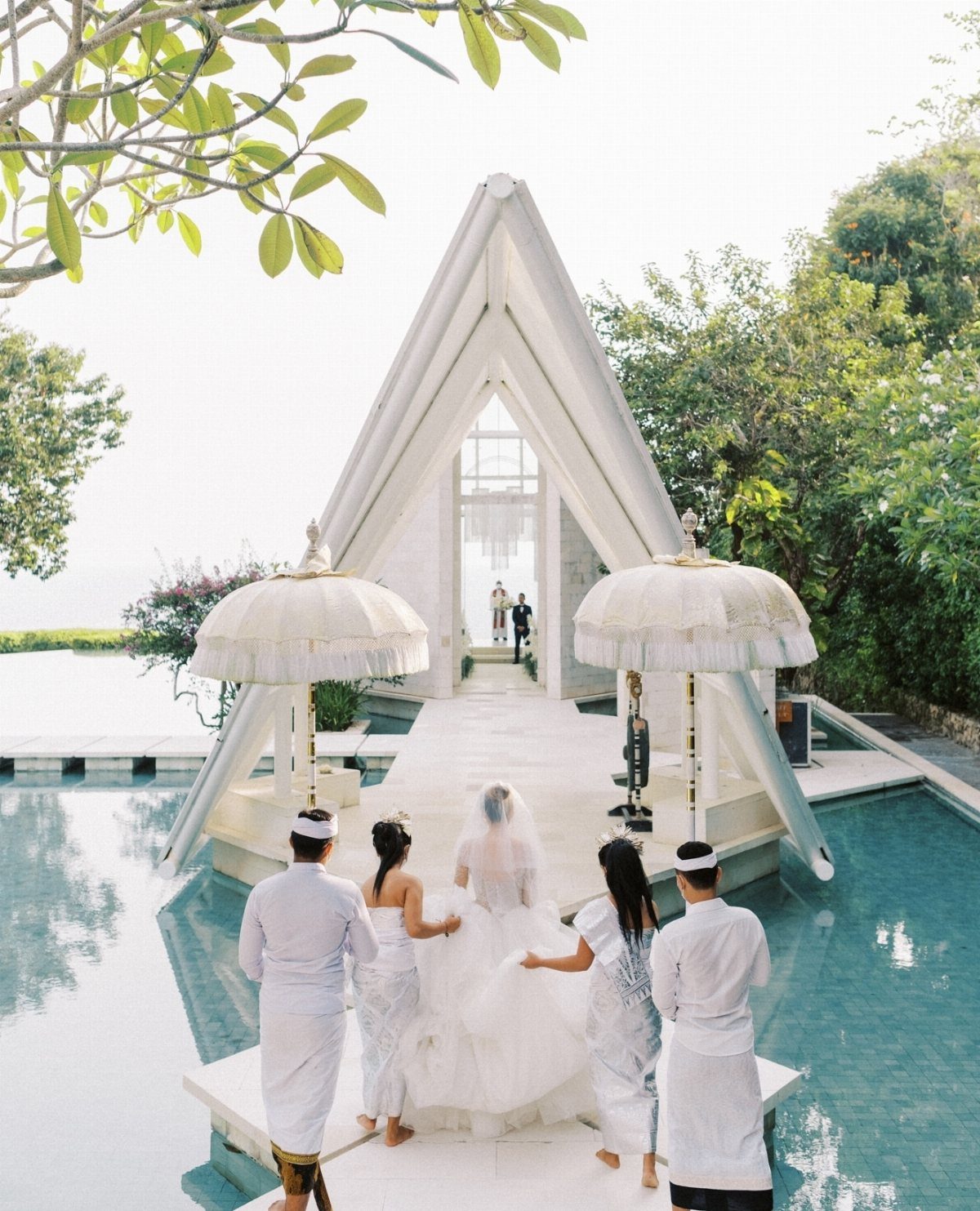 Bride walking toward a white A-frame glass chapel surrounded by reflective water pools.