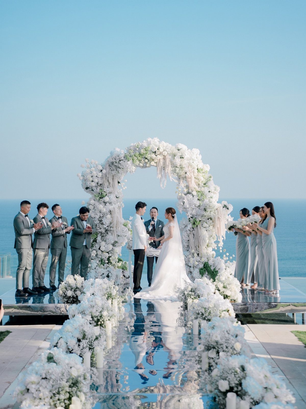 Wedding couple under a white floral arch on a mirrored aisle overlooking the ocean