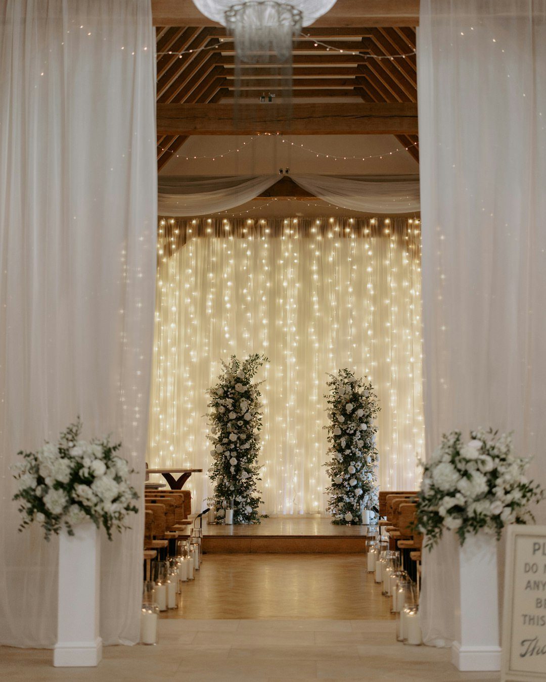 Wedding ceremony backdrop with fairy lights, white curtains, and white floral arrangements.