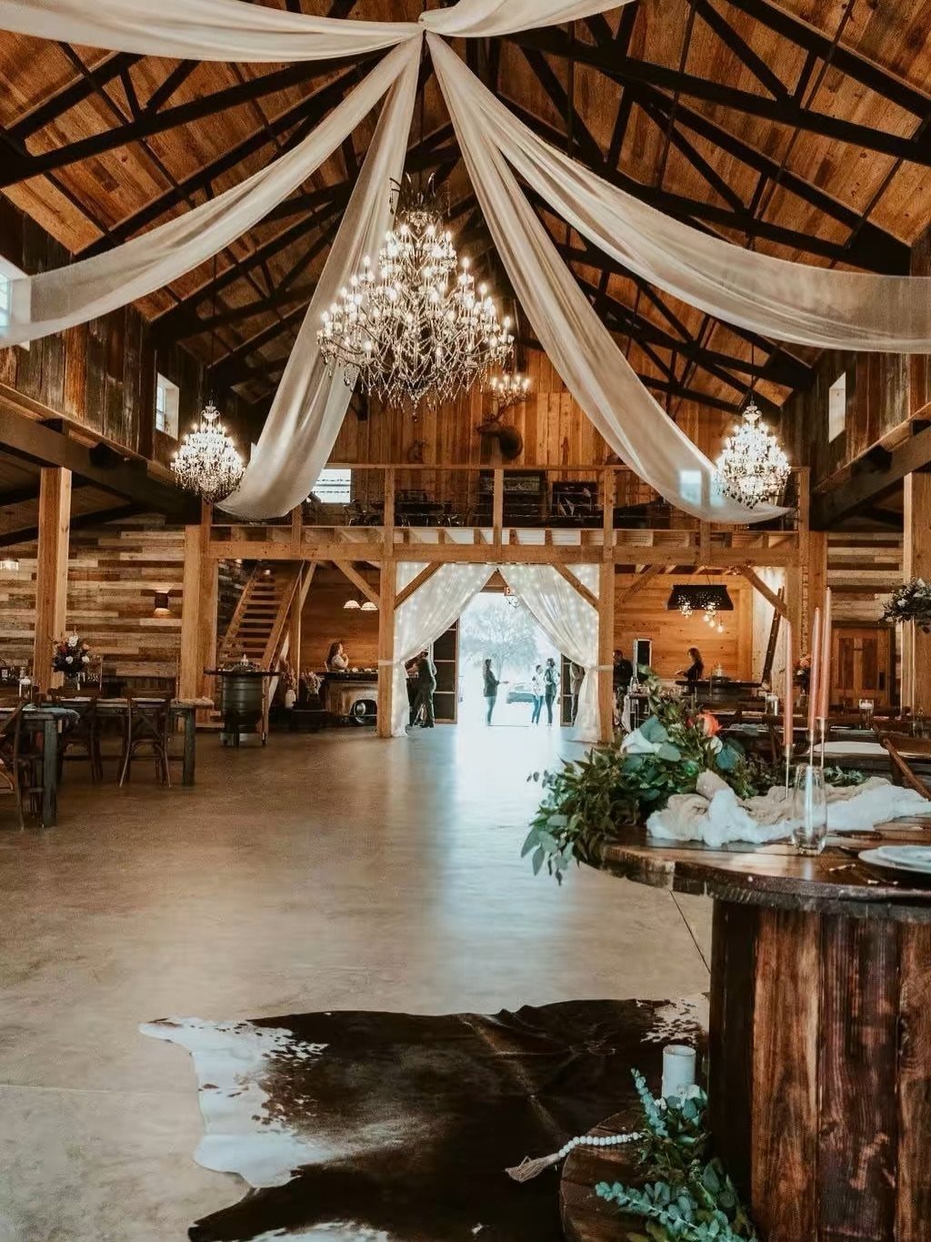 Rustic barn interior with white ceiling drapes, crystal chandeliers, and wooden wedding tables.