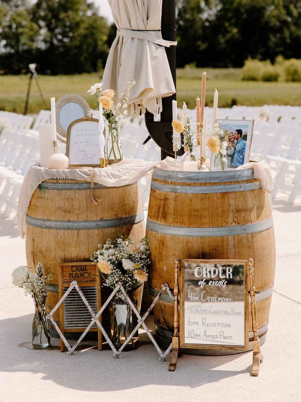 Wooden barrels decorated with flowers, candles, and a wedding schedule sign outdoors.