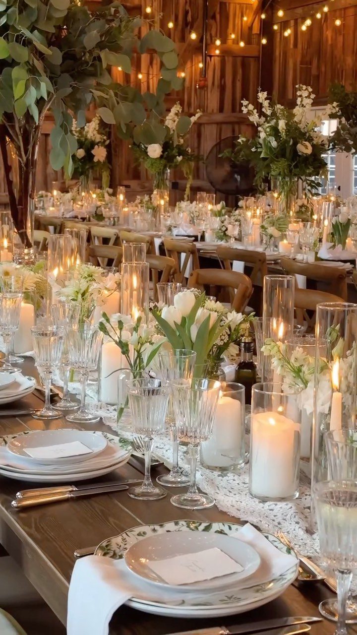 Long wooden reception table with white flowers and candles inside a rustic barn
