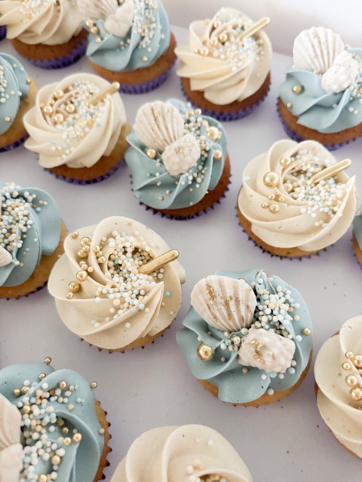 Cupcakes with blue and cream frosting decorated with seashells and gold pearls.
