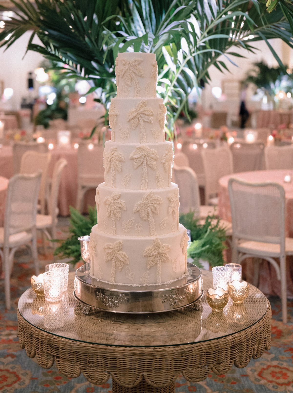 Five-tier white wedding cake with palm tree designs on a wicker table.