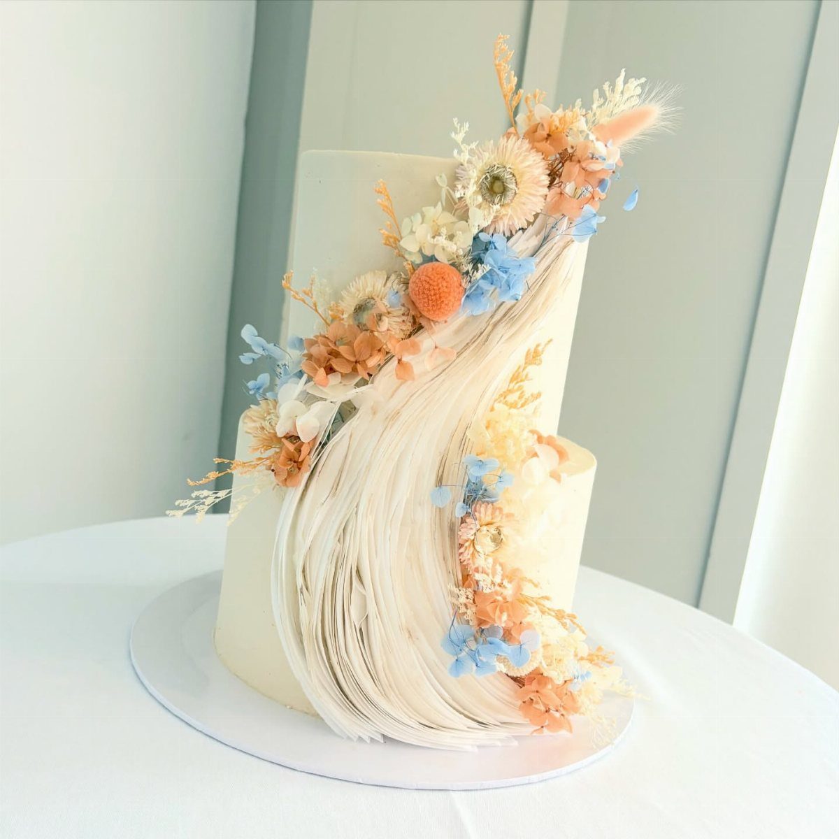 Two-tier white cake with a frosting wave and blue and orange floral decorations.