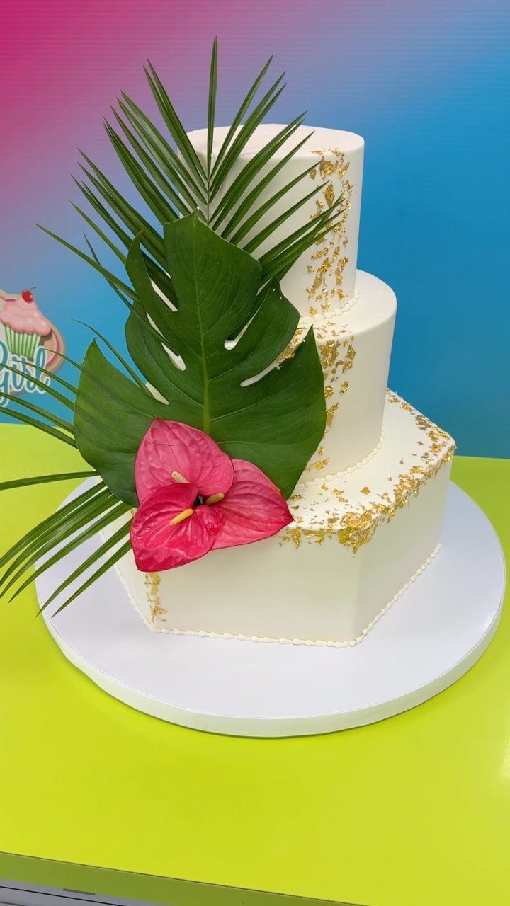 Three-tiered white cake with gold leaf, green tropical leaves, and a pink anthurium flower.