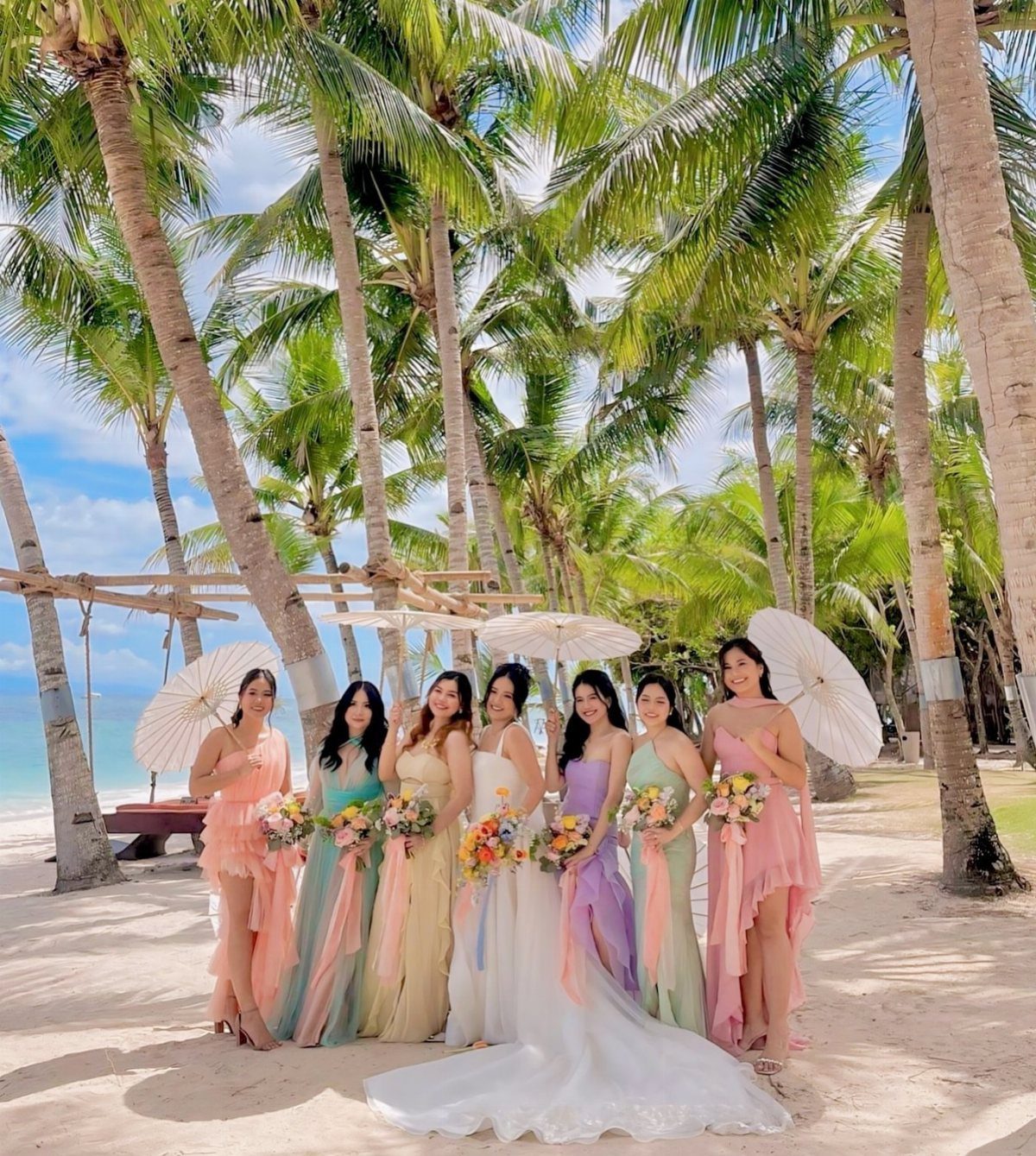 Bride and bridesmaids in pastel dresses posing on a tropical beach with palm trees.