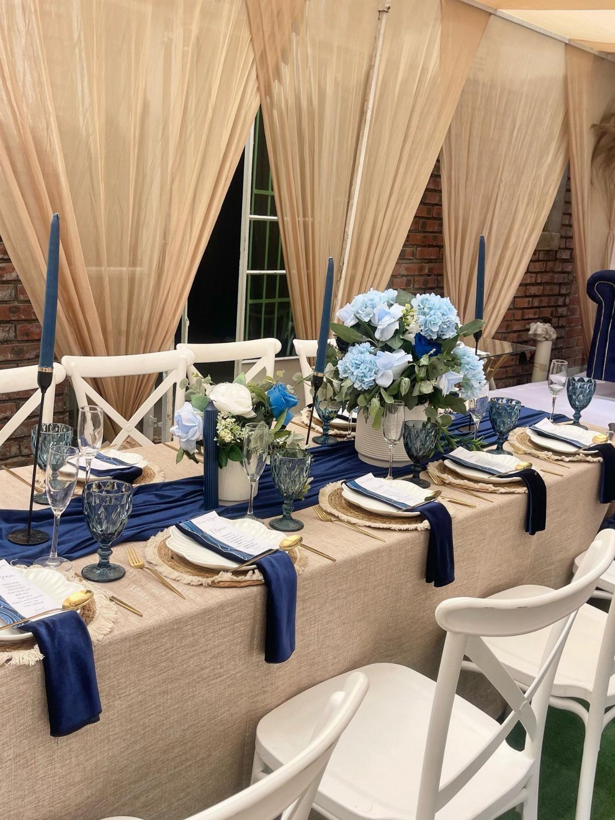 Wedding table setting with navy blue accents, beige linens, and blue floral centerpieces.