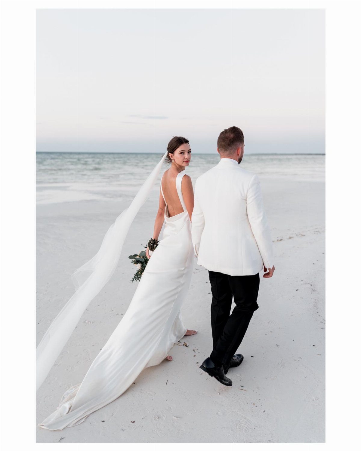 Bride and groom walking on a white sand beach toward the ocean waves.