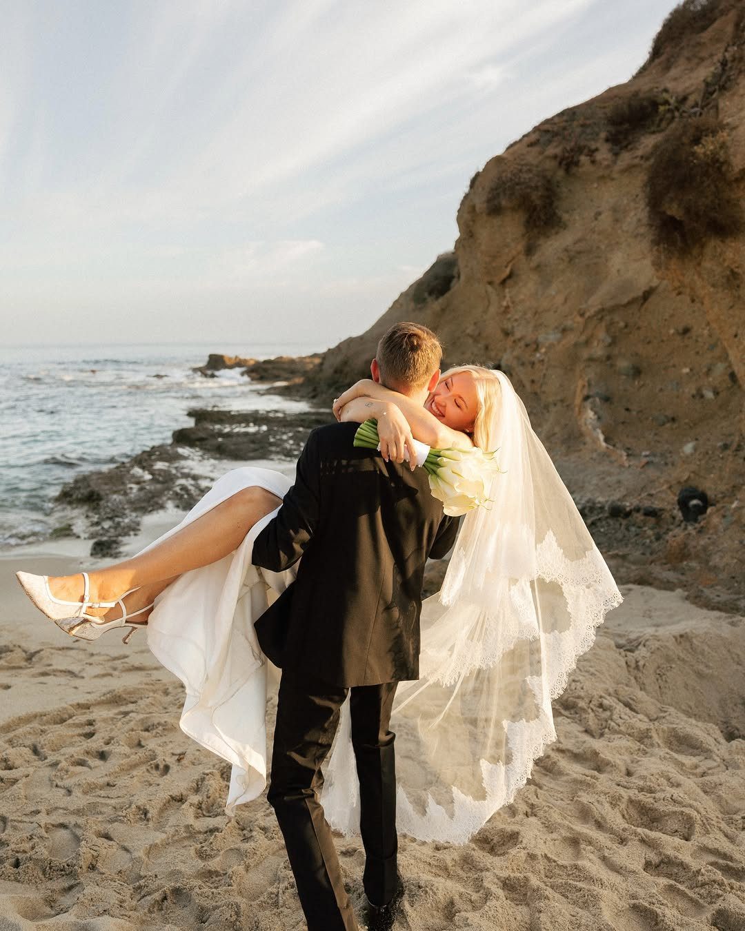 Groom carrying bride in wedding attire on a sandy beach by the ocean