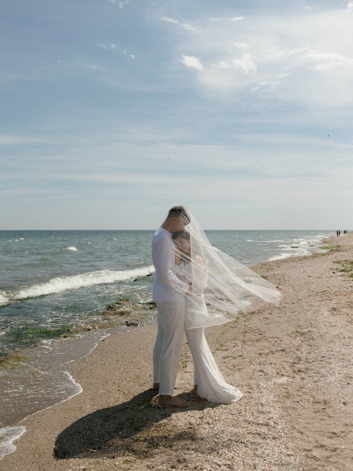 Couple embracing on a beach with a long white veil blowing in the wind.