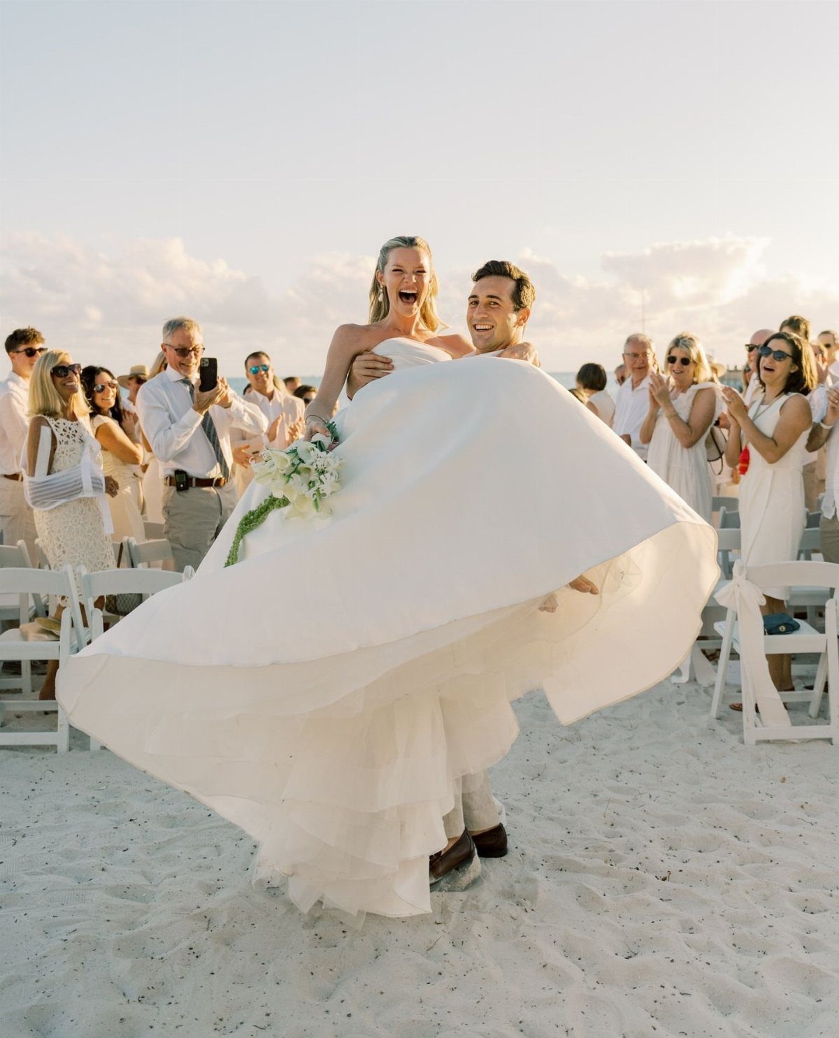 Groom carrying laughing bride in white dress on a sandy beach with guests.