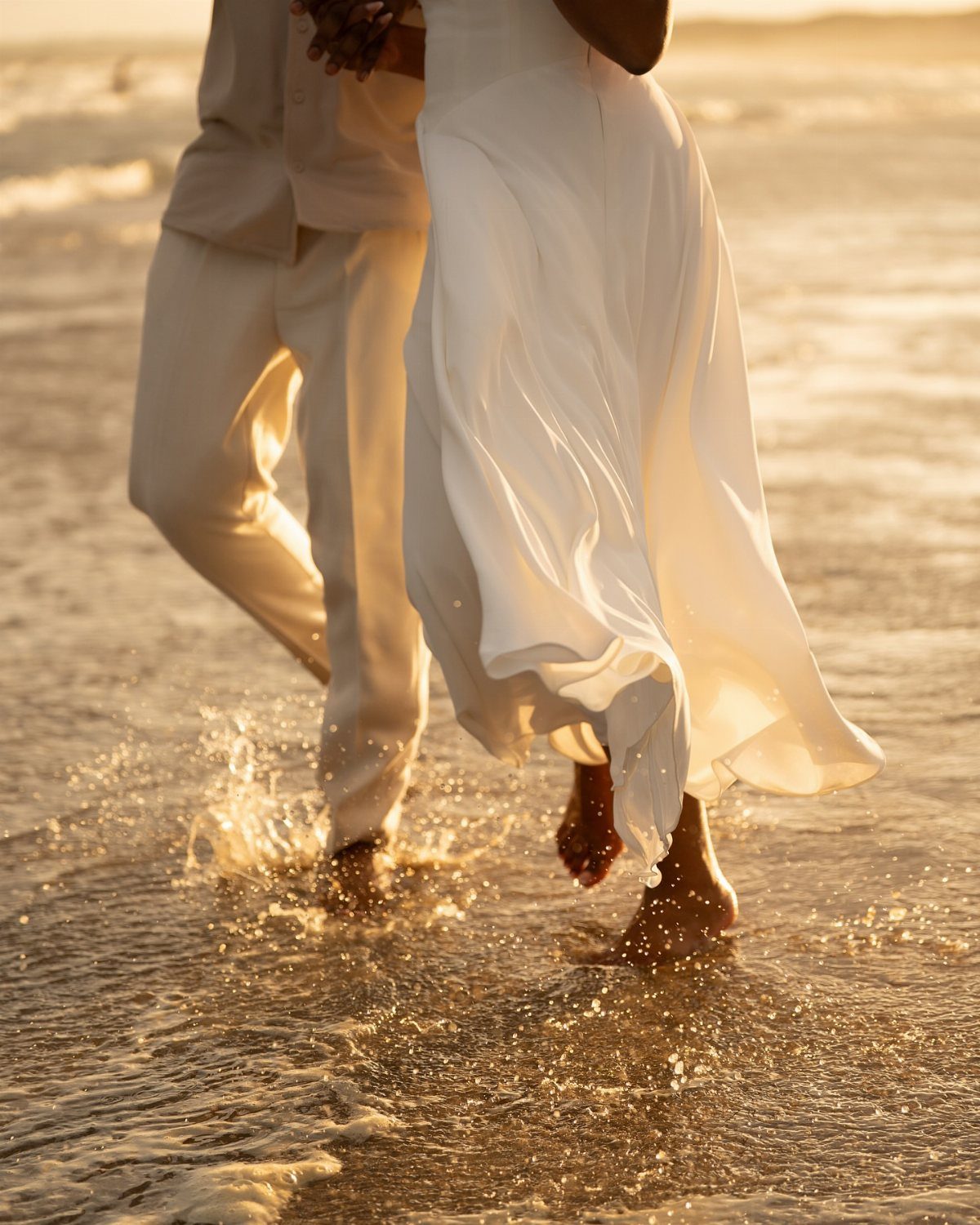 Wedding couple walking barefoot through shallow ocean water during a golden sunset.