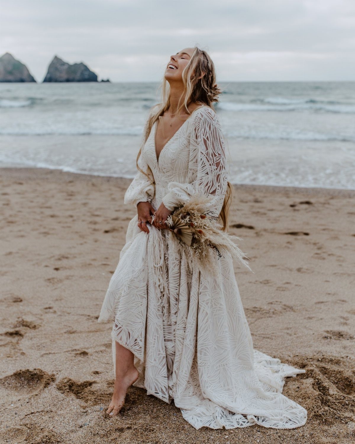 Woman in a flowy white lace wedding dress smiling on a sandy beach