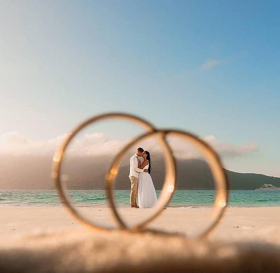 Gold wedding rings framing a bride and groom kissing on a tropical beach.