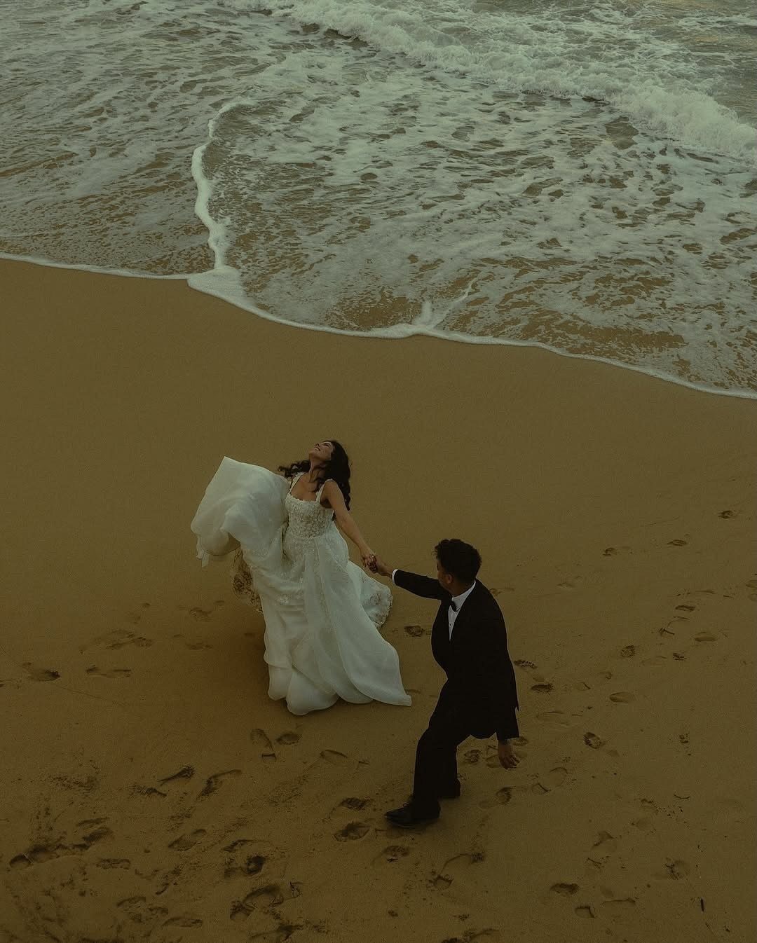 Bride and groom holding hands while walking on a sandy beach by the ocean