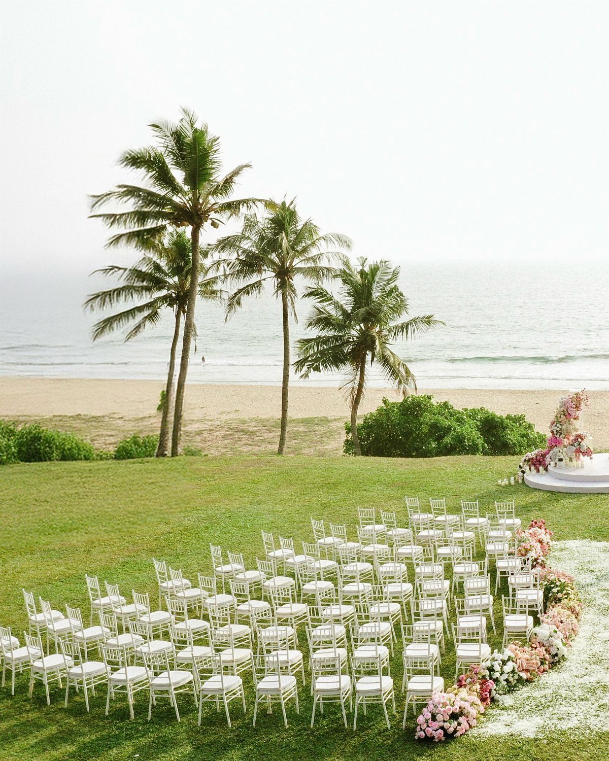 White wedding chairs on a green lawn overlooking a beach with palm trees.
