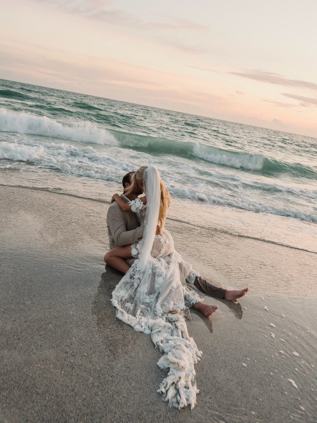 Bride and groom embracing and kissing on a sandy beach with crashing waves.