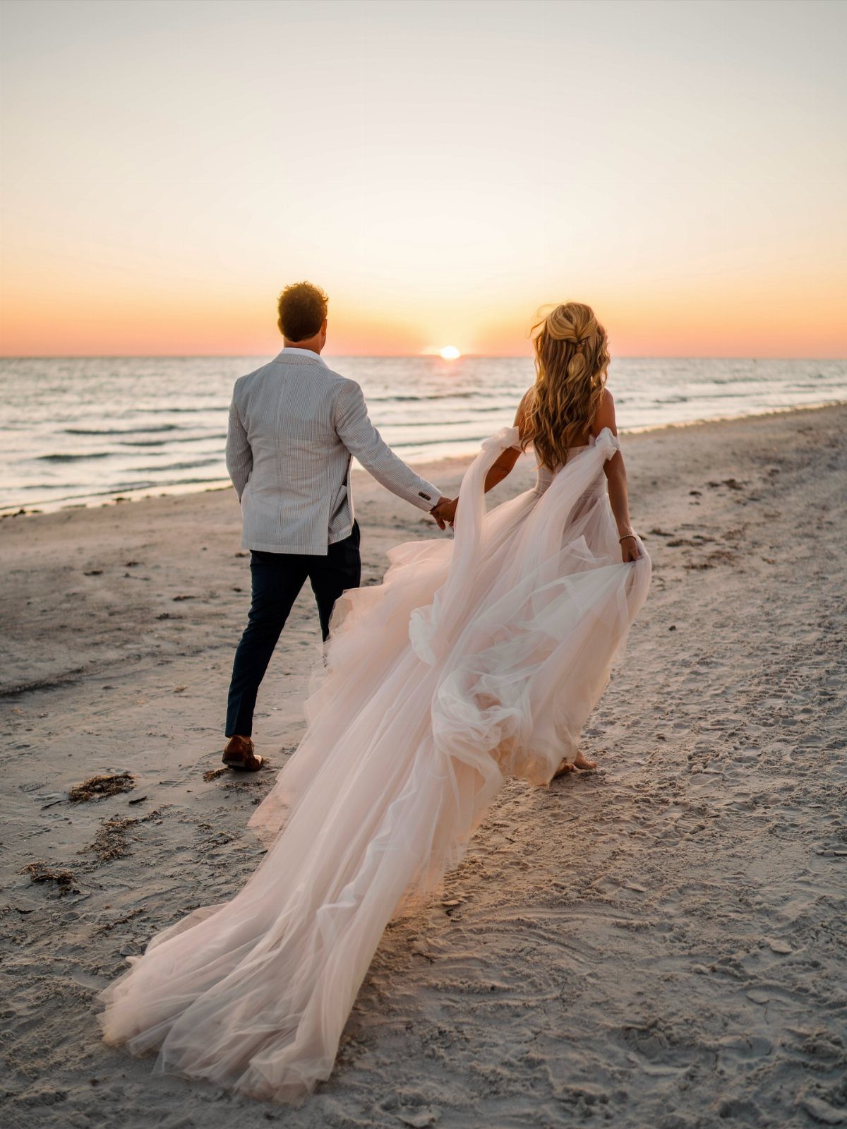 Bride and groom walking hand-in-hand on a beach during a golden sunset.