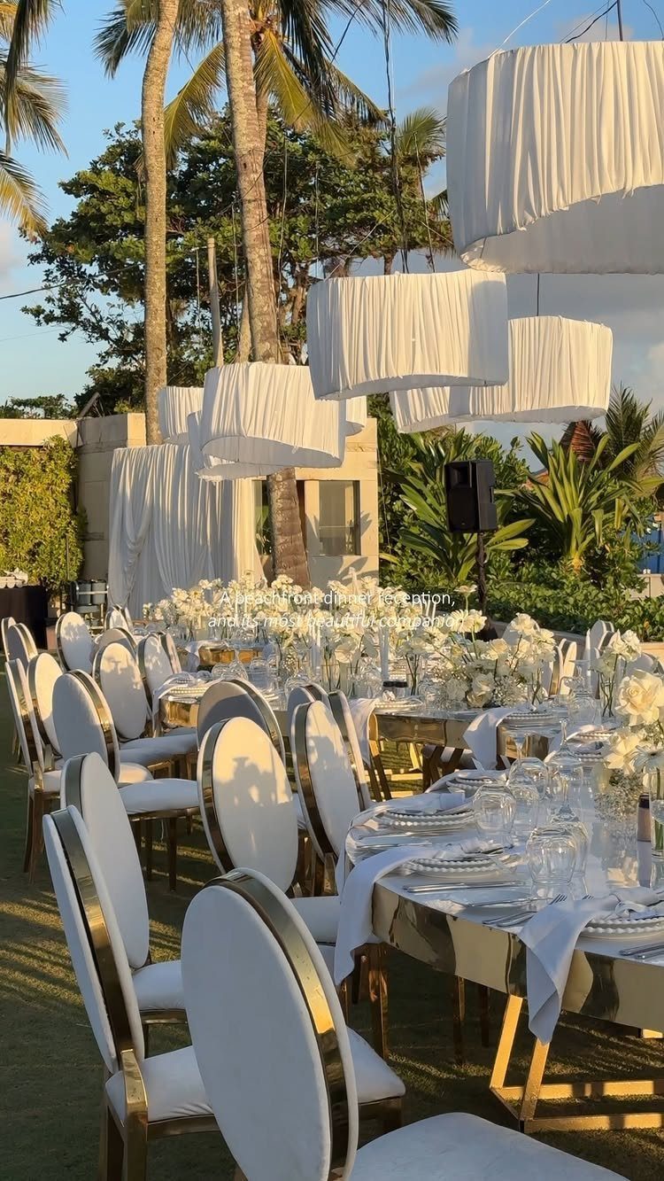 Elegant outdoor wedding reception table with white flowers, gold chairs, and hanging lampshades.