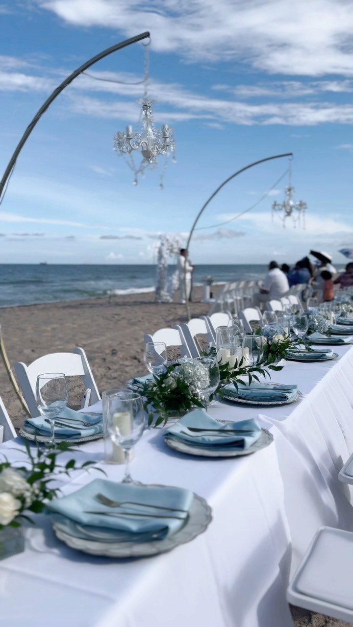 Long white dining table with blue napkins and chandeliers on a sandy beach