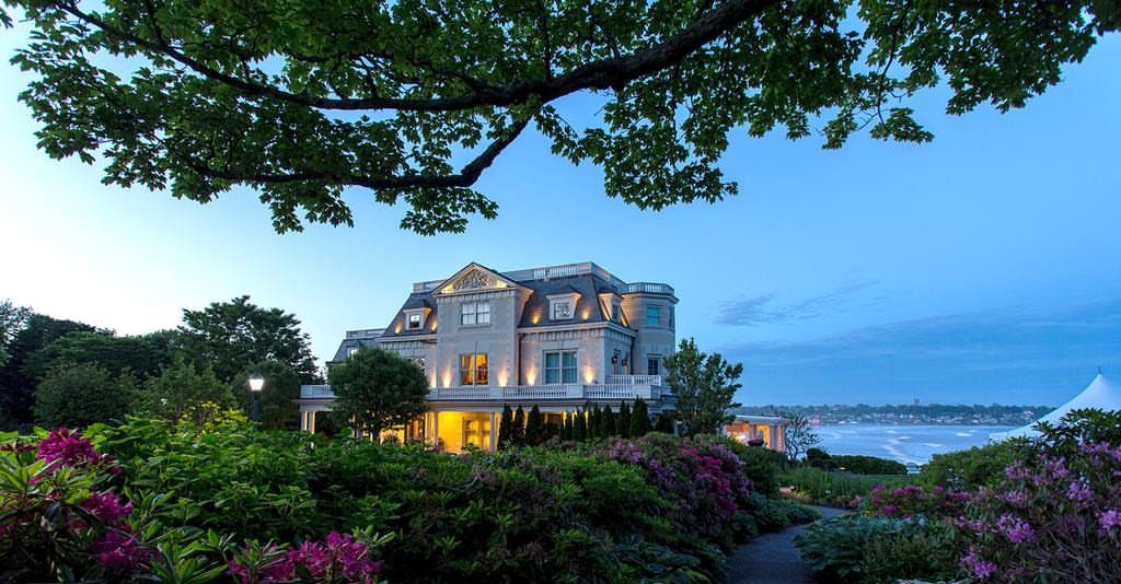 Illuminated stone mansion with ocean views surrounded by lush gardens and purple flowers.