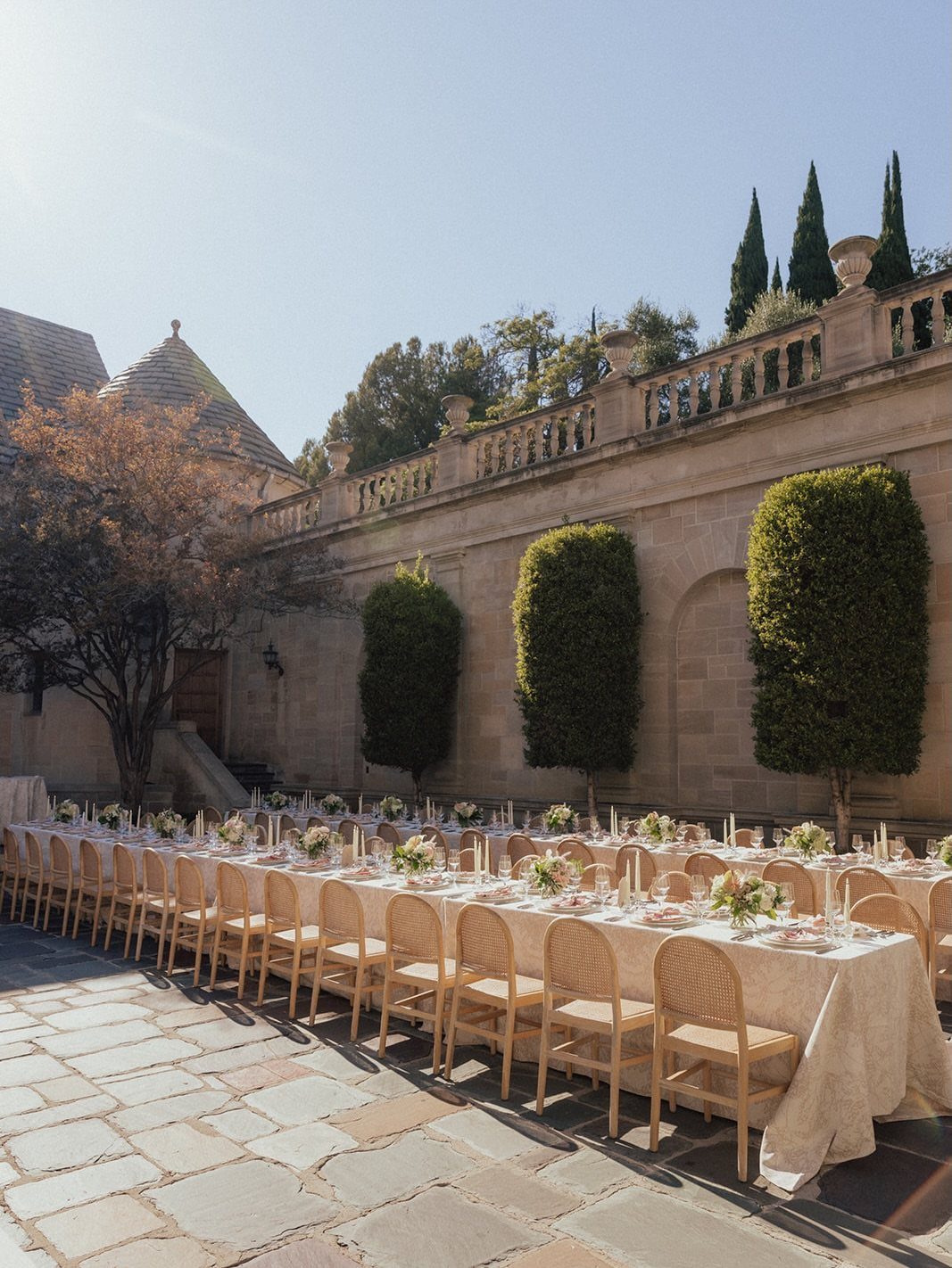 Long banquet table with gold chairs on a stone patio at a stately mansion.