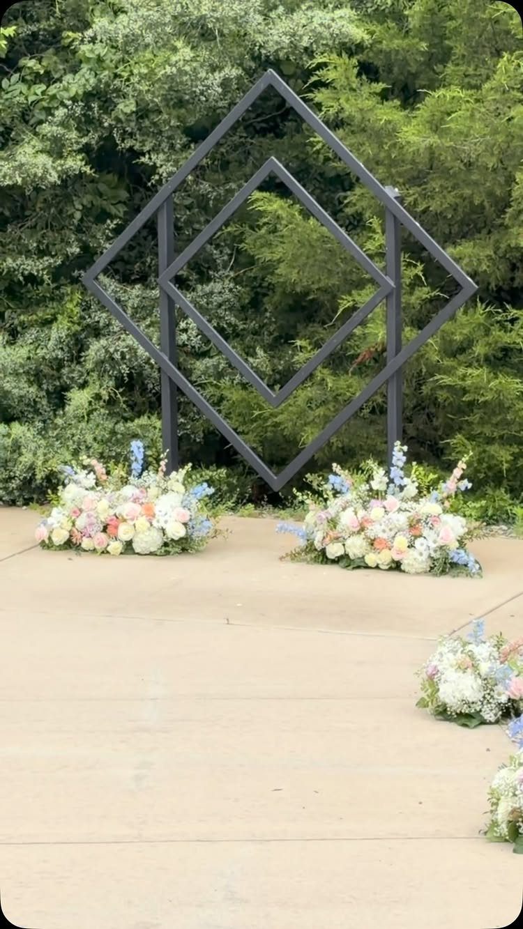 Black geometric diamond wedding arch with pastel flowers against a green forest backdrop