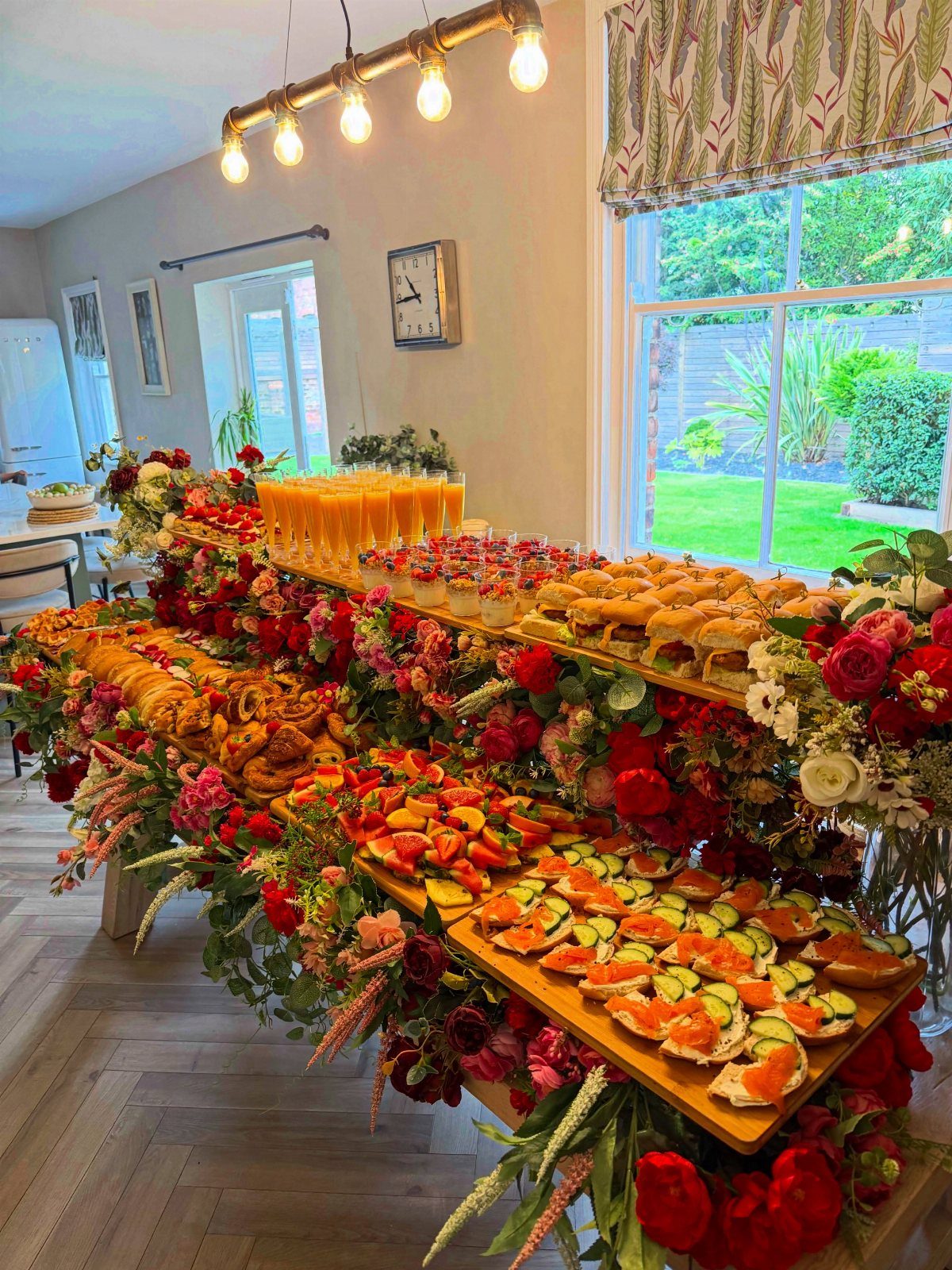 Tiered food display with red roses, appetizers, and orange juice by a window.