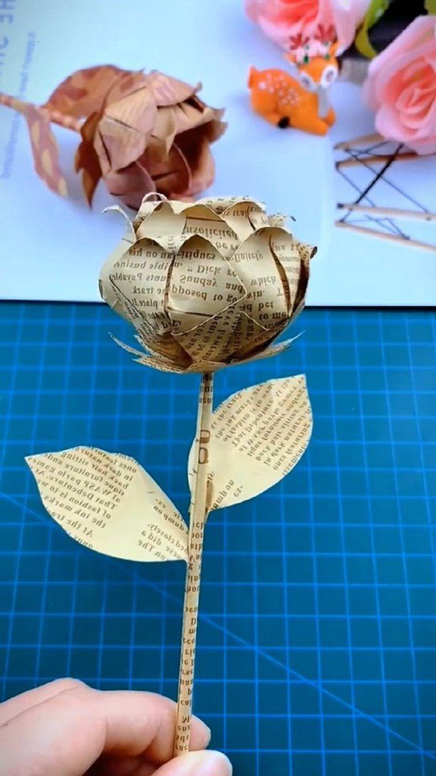 Handmade rose crafted from vintage text-printed paper held against a blue gridded mat.