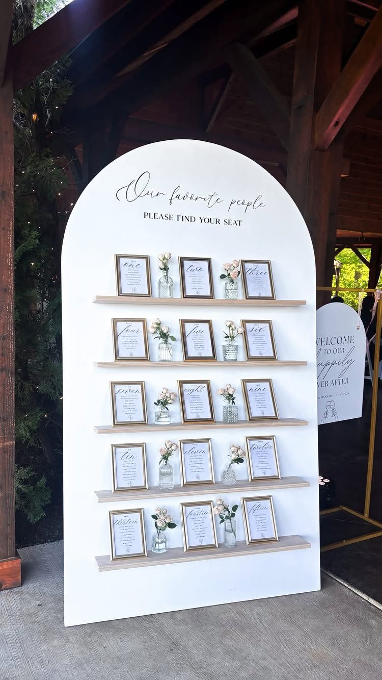 White arched seating chart with wooden shelves, gold frames, and small white flowers.