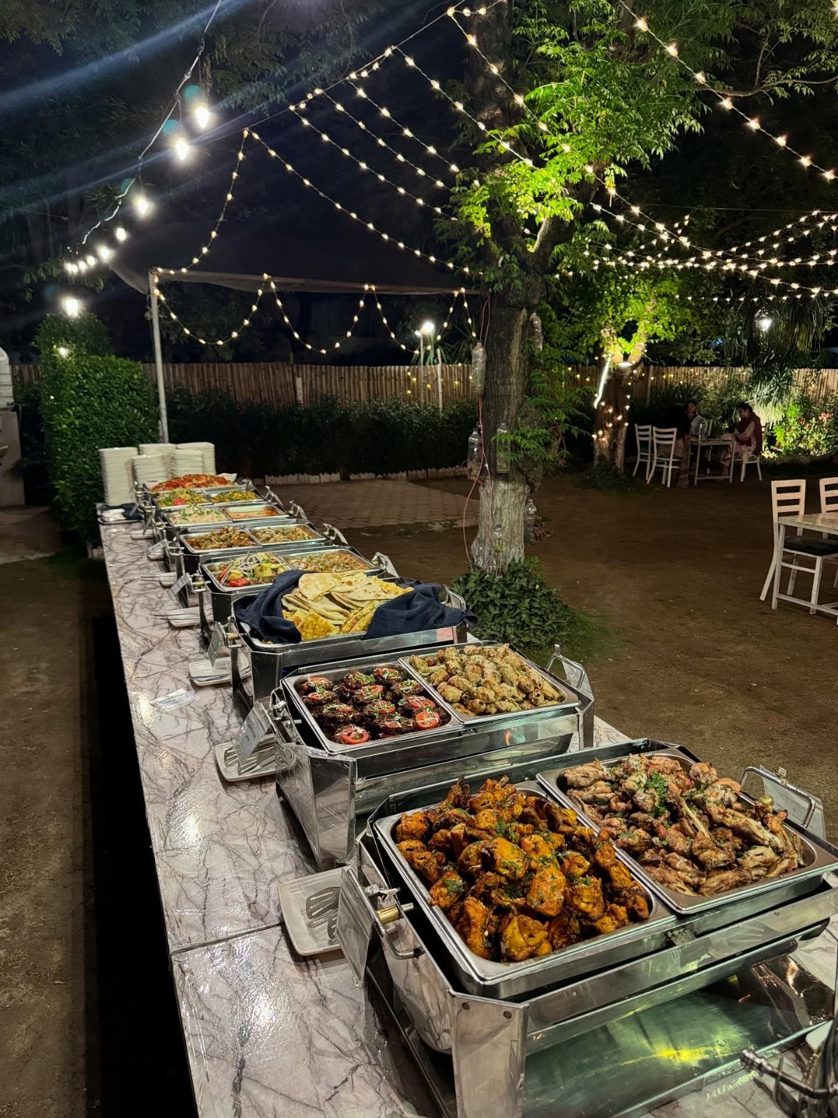 Outdoor buffet table with silver chafing dishes of grilled meats under string lights.