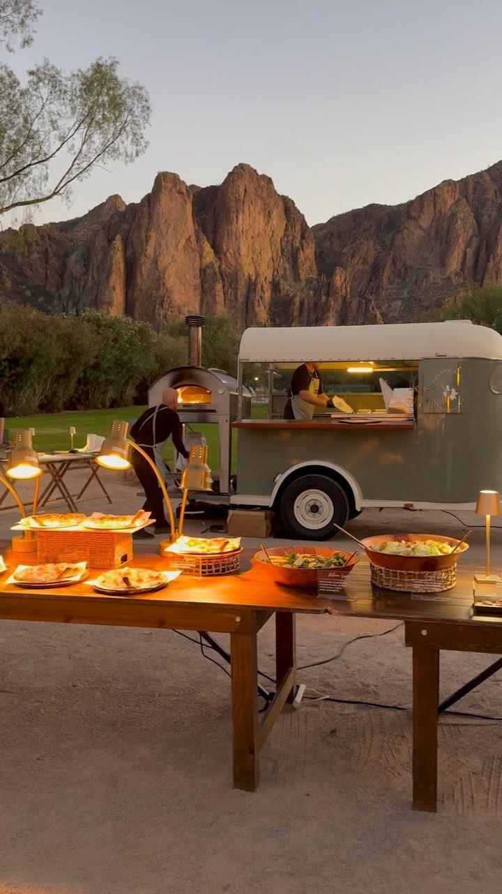 Vintage grey food truck with pizza oven and buffet tables against mountain backdrop