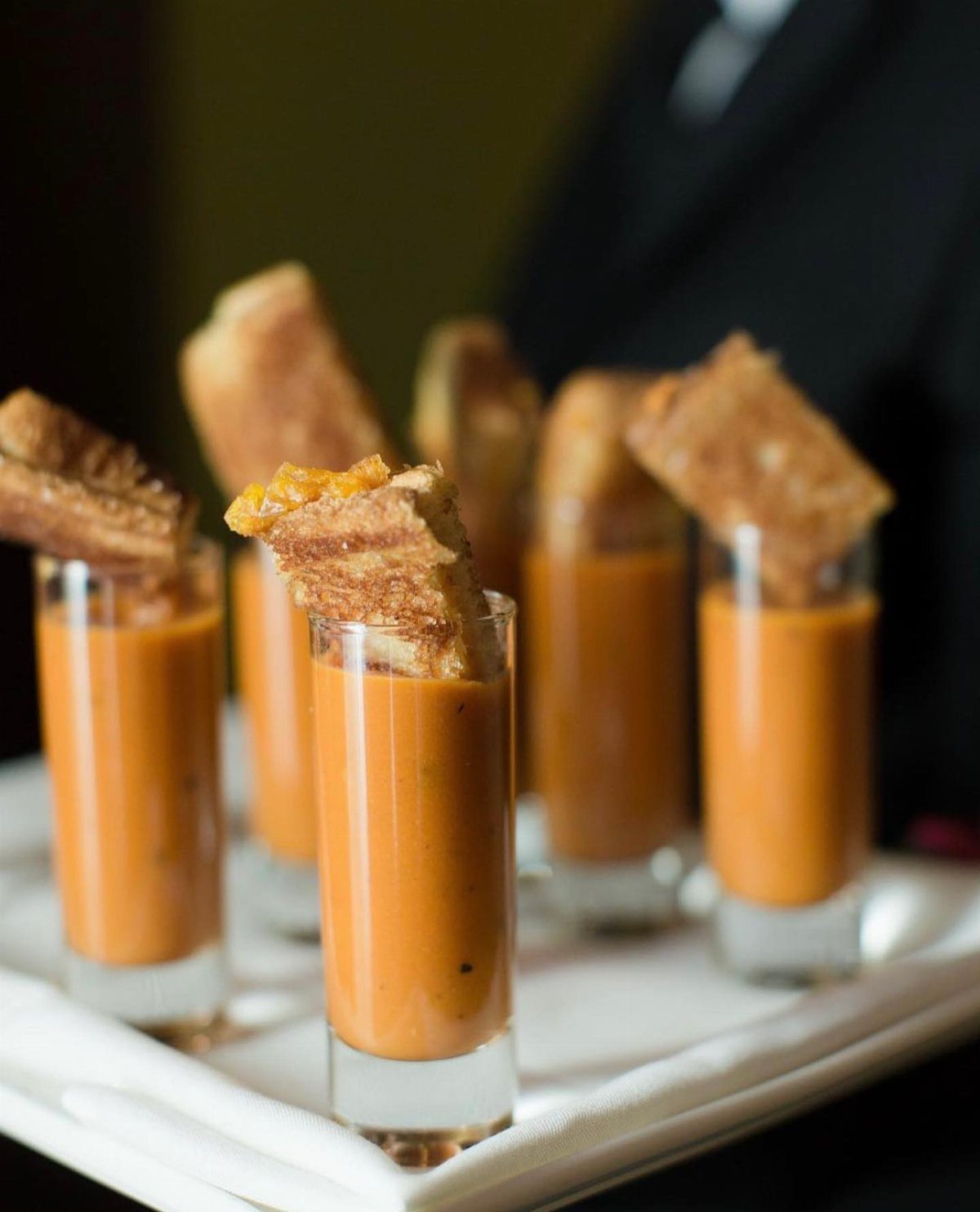 Tomato soup shooters topped with small toasted grilled cheese sandwich wedges on white napkin