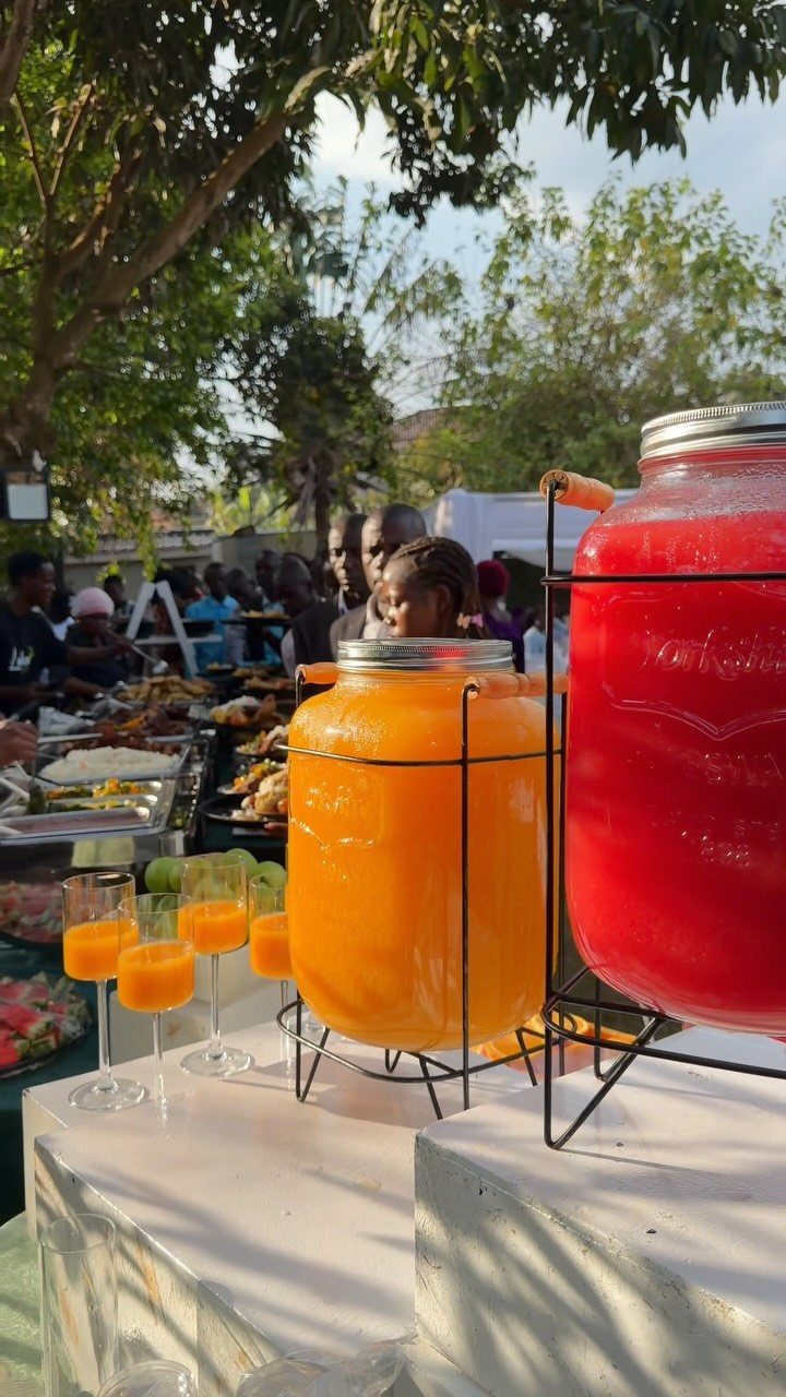 Beverage dispensers with orange and red drinks and a buffet in the background.