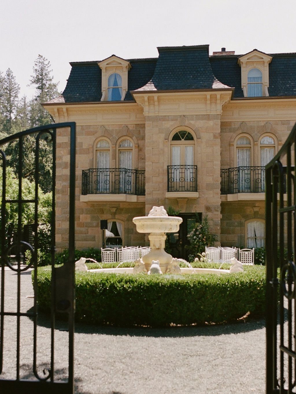 Stone manor house with a tiered fountain and manicured hedges behind open iron gates.