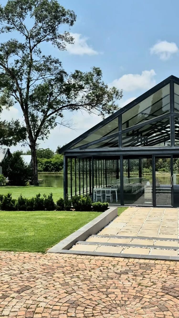 Modern glass pavilion wedding venue with black frames, green lawn, and cobblestone path.