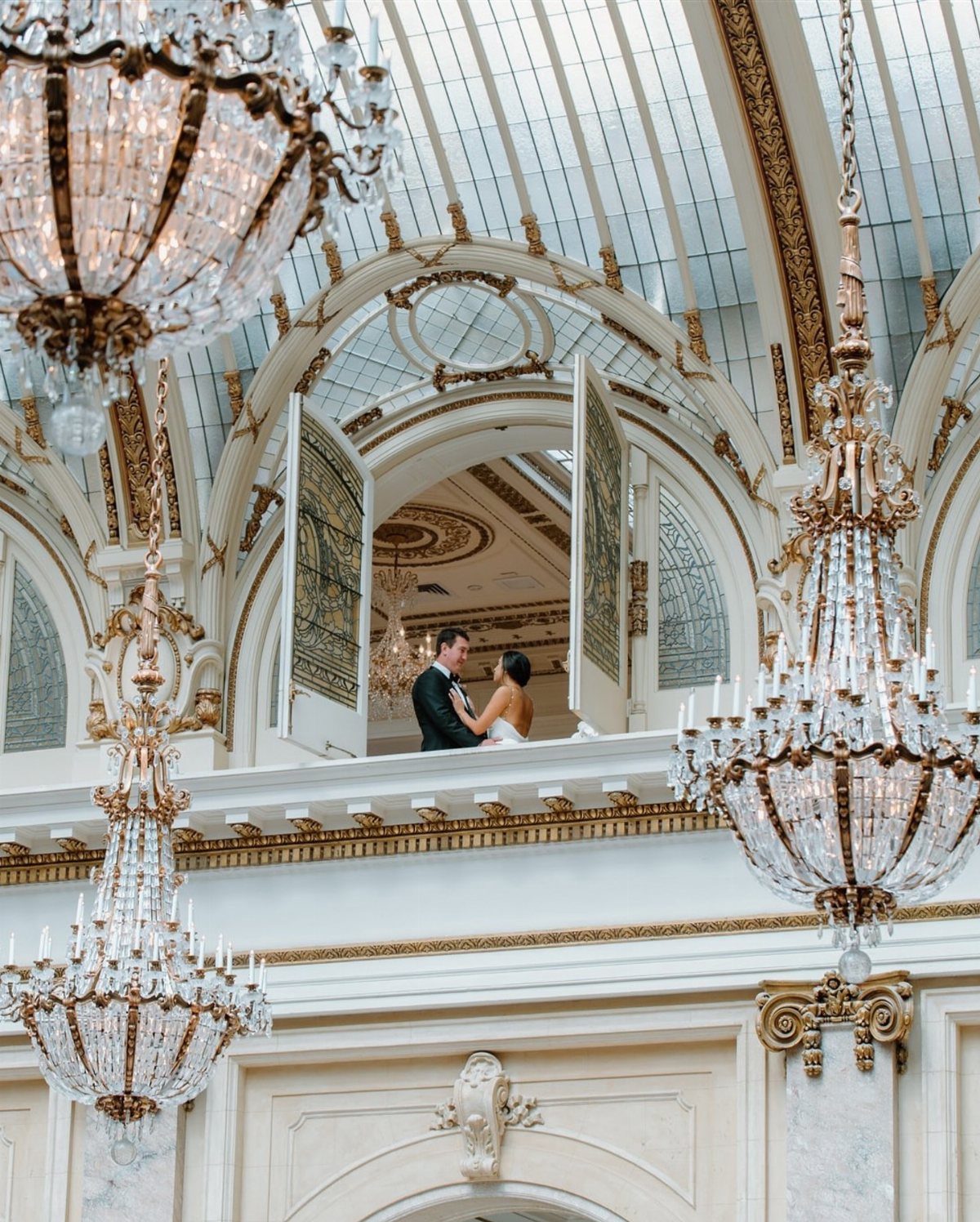 Bride and groom embracing on a balcony under crystal chandeliers in an ornate hall
