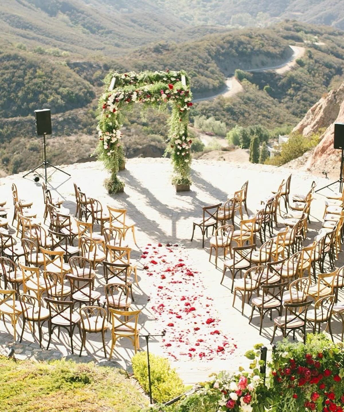 Outdoor wedding ceremony setup with wooden chairs, floral arch, and red petal aisle.