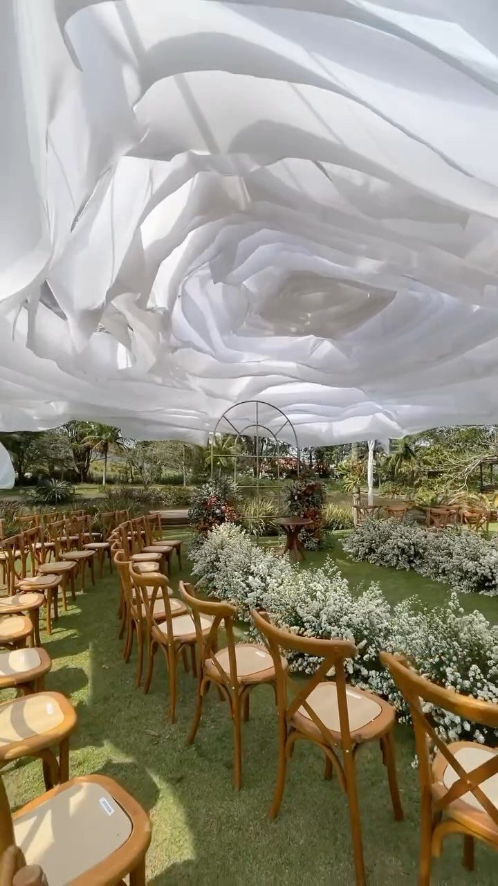 Outdoor ceremony setup with wooden chairs, white floral aisle, and draped white fabric canopy.