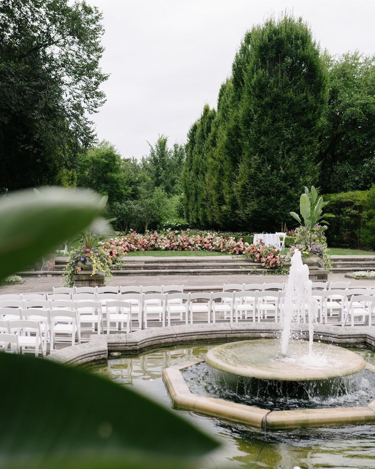 White ceremony chairs and a floral arch next to a stone water fountain.