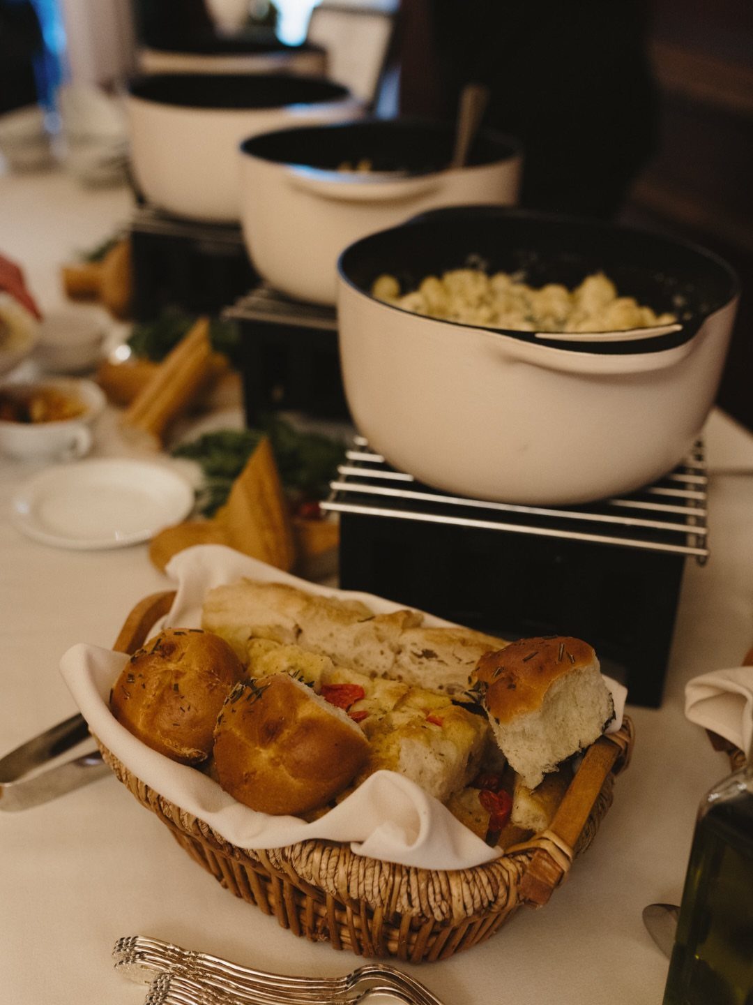 Assorted bread rolls in a basket next to white buffet warming pots.