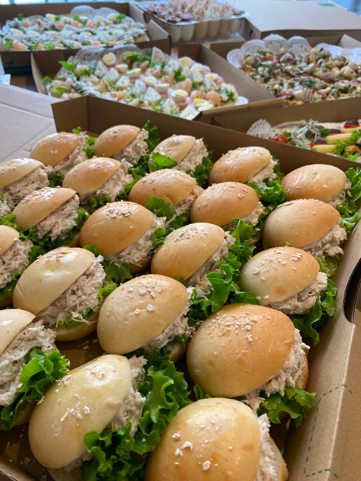 Small slider sandwiches with creamy filling and lettuce in cardboard catering boxes.