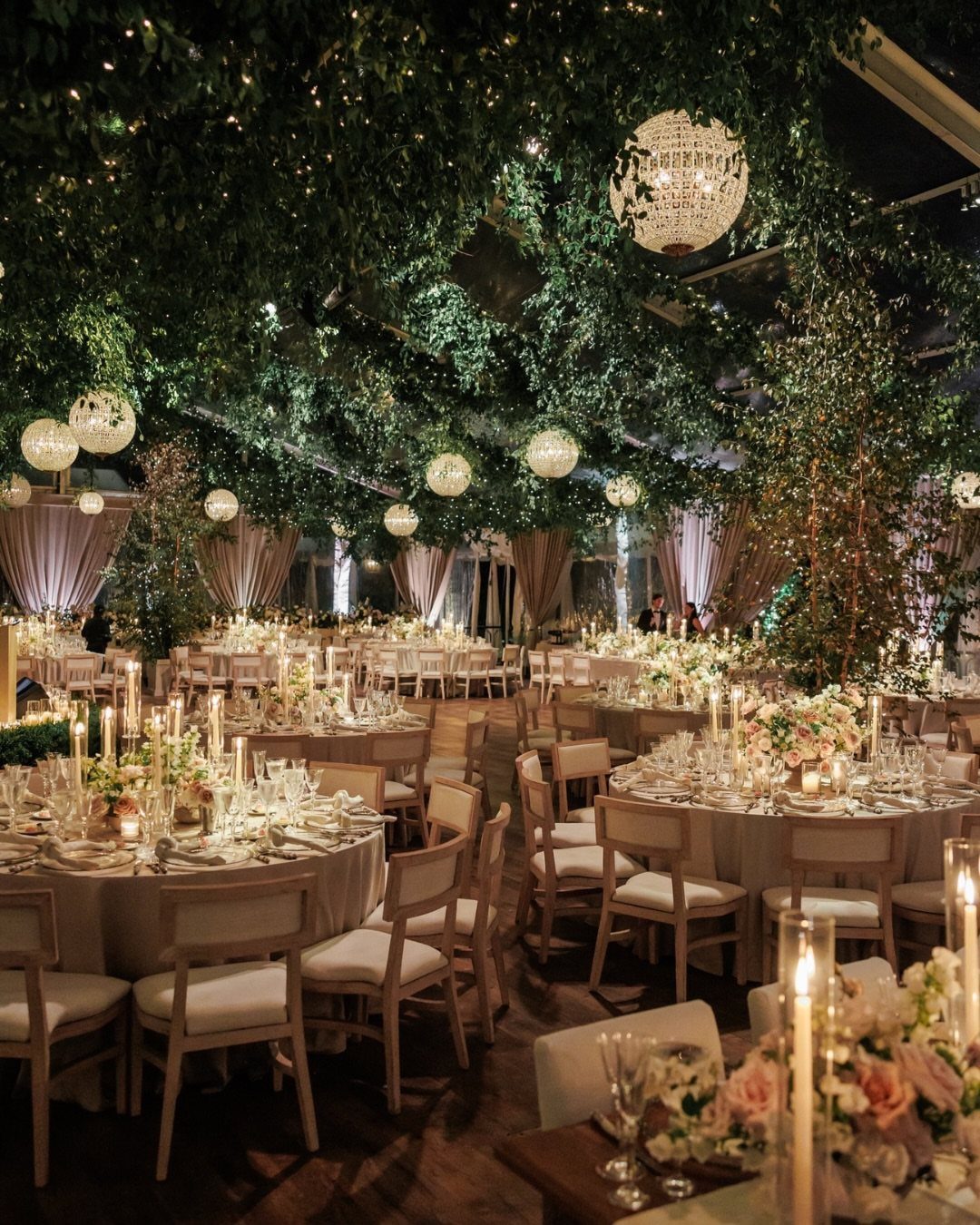 Wedding reception tables under a ceiling of lush greenery and crystal chandeliers.