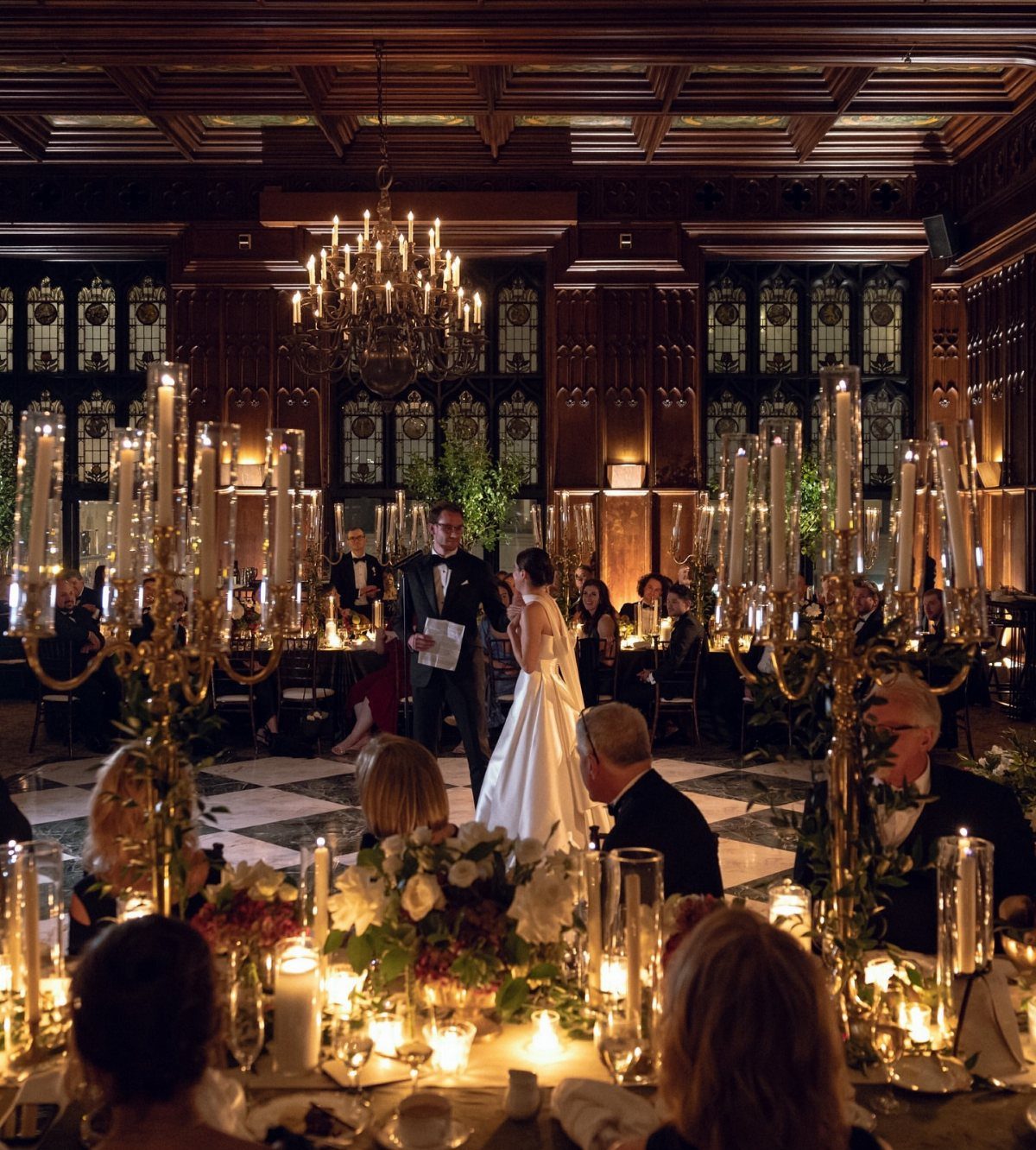 Groom speaking to bride in a dark wood-paneled historic room with gold candelabras.