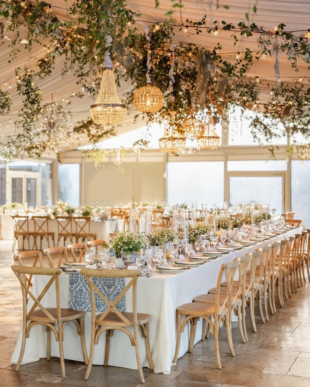Wedding banquet table under a white tent with hanging greenery and crystal chandeliers