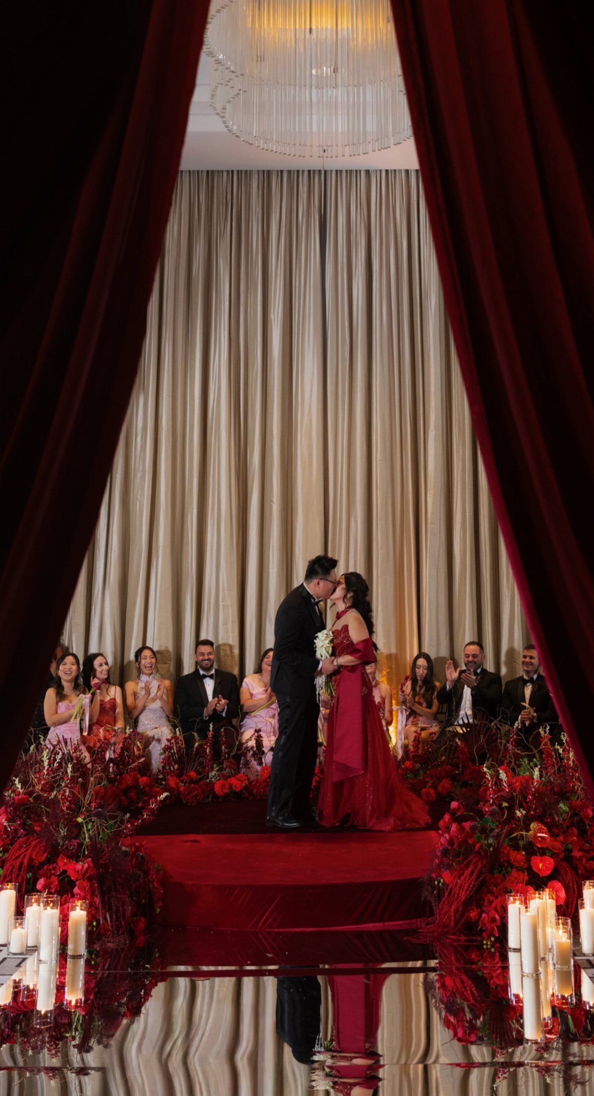 Couple kissing on a red platform surrounded by red flowers and formal guests.