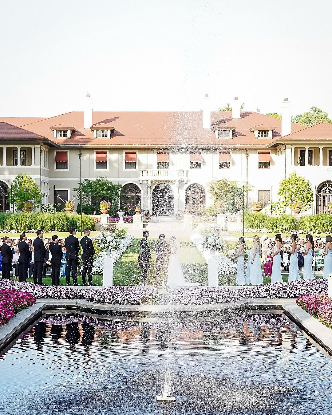 Wedding ceremony outdoors in front of a large estate with a reflecting pool.