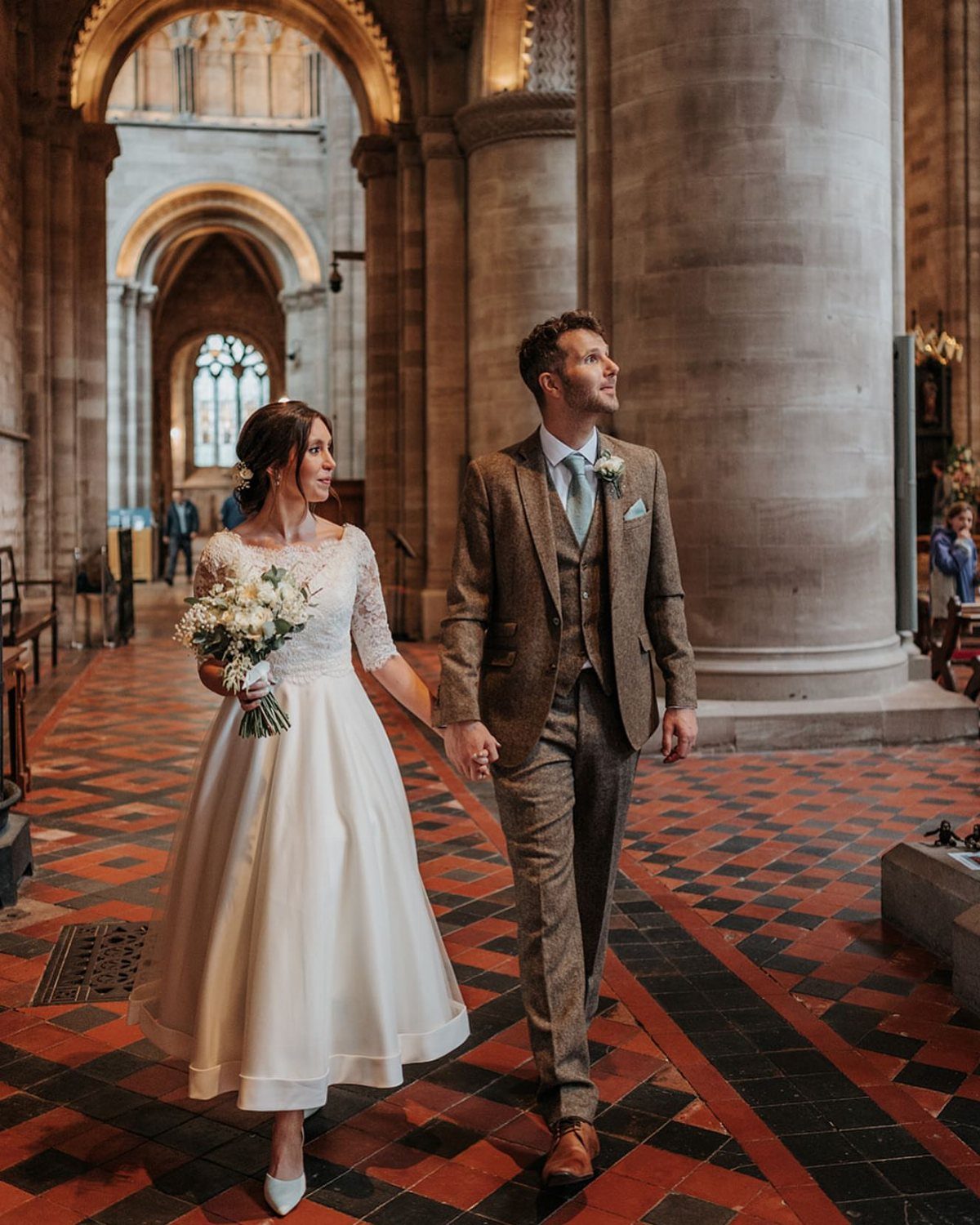 Bride in knee-length white bridal gown and groom in brown tweed suit walking together.