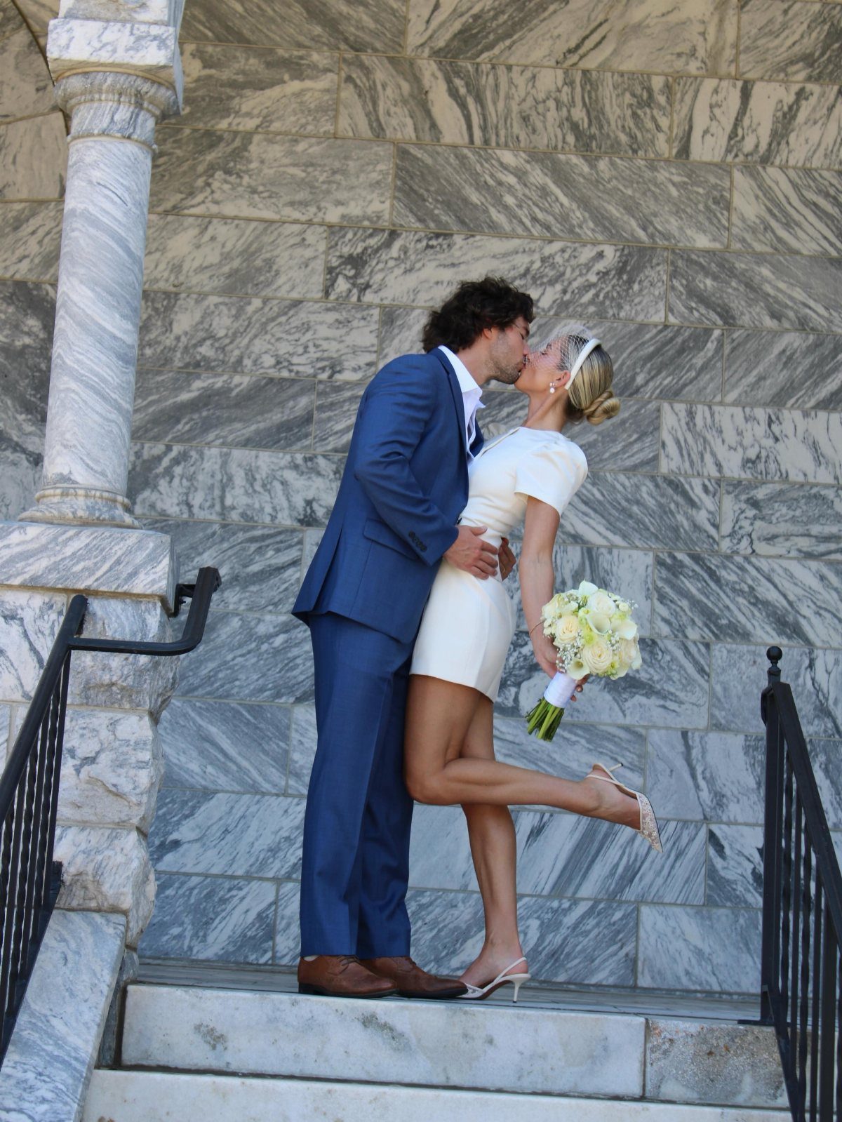 Couple kissing on marble steps, woman wearing a short white wedding dress and heels.